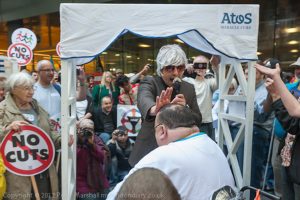 The 'Atos Miracle Cure' -'Atos's own Reverend' blesses and lays his hand on the head of a man in a wheelchair at the Closing Atos Ceremony by DPAC & UK Uncut outside Atos's offices.