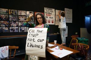Woman standing in front of an exhibition. She is holding a poster which says "Cut your crap not our lifelines"