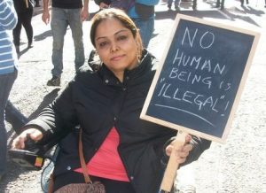 Photo of Manjeet Kaur wearing a black jacket sitting in her wheelchair holding a placard saying 'NO human is "illegal"!'