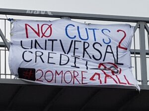 A photo of a white banner hanging from a bridge saying "No Cuts 2 Universal Credit #20More4All"