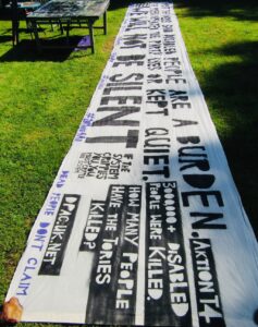 Photo of a long black and white banner laid out on grass stretching out into the distance. One of the slogans on it says "WE WILL NOT BE SILENT"
