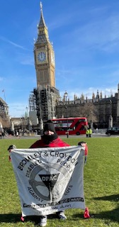 DPAC member Paula Peters standing in Parliament square holding a black and white banner that says Bromley and Croydon Disabled People Against Cuts Banner at Parliament Square supporting the London Cost of living demo in London called by DPAC, Peace and Justice Campaign, Revolutionary Socialist Group 21, Fuel Poverty Action and Peoples Assembly