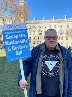 DPAC member Peter Rogers wearing a black and white dpac t shirt holding a placard in his left hand that says Scrap the Nationality and Borders Bill was standing at Parliament square waiting for the London Cost of Living Demo to begin