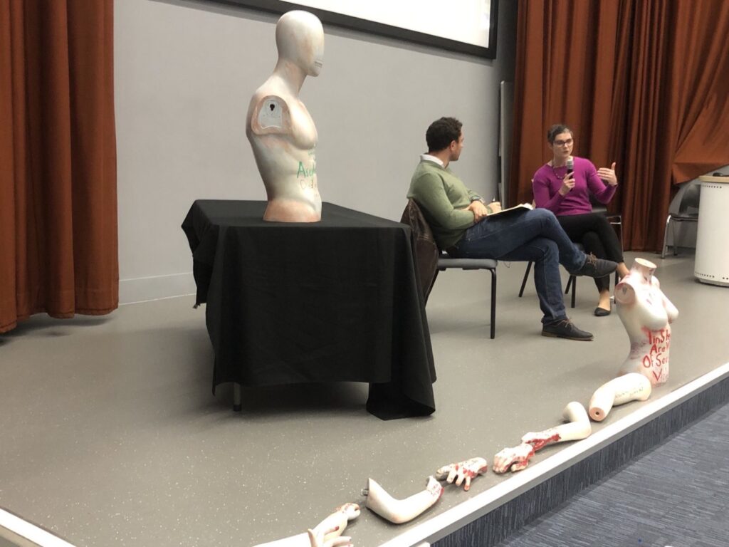 Chloe Smith in a pink top sitting on the lecture theatre stage, being interviewed by a man in a green top. In the foreground is an anatomical bust of a man on a table. Along the front of the stage are body parts from an anatomy dummy. (I'm not sure if that was part of the lecture or protest!)