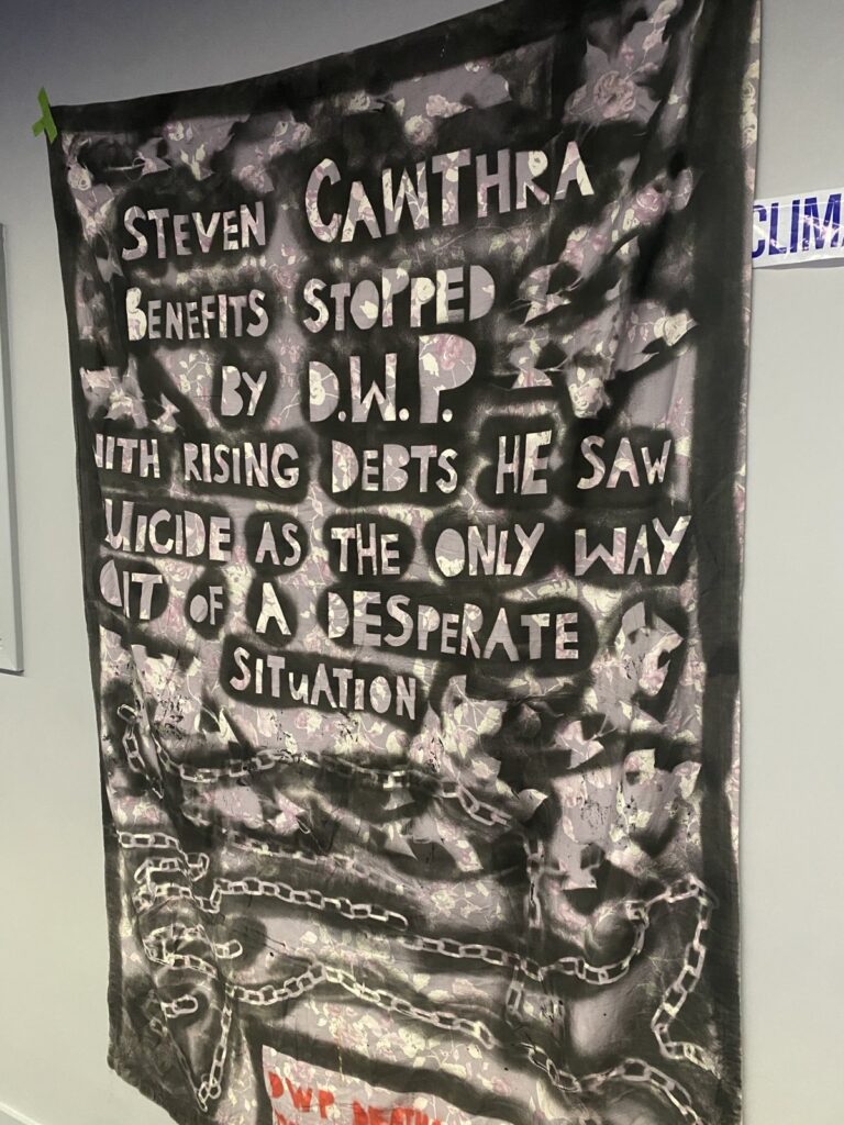 One of the 'DWP Deaths Make Me Sick' shrouds taped to the wall. Spray painted text on the shroud reads: Steven Cawthra Benefits stopped by DWP with rising debts he saw suicide as the only way out of a desperate situation. ‘DWP Deaths Make Me Sick.’