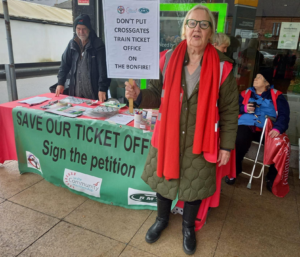 Margaret Batty holding a placard saying “Dont put Cross Gates train ticket office on the bonfire”; in front of table with banner showing DPAC, Unite Community, and RMT logos, table with handouts and petitions staffed by DPAC and unite Community members;