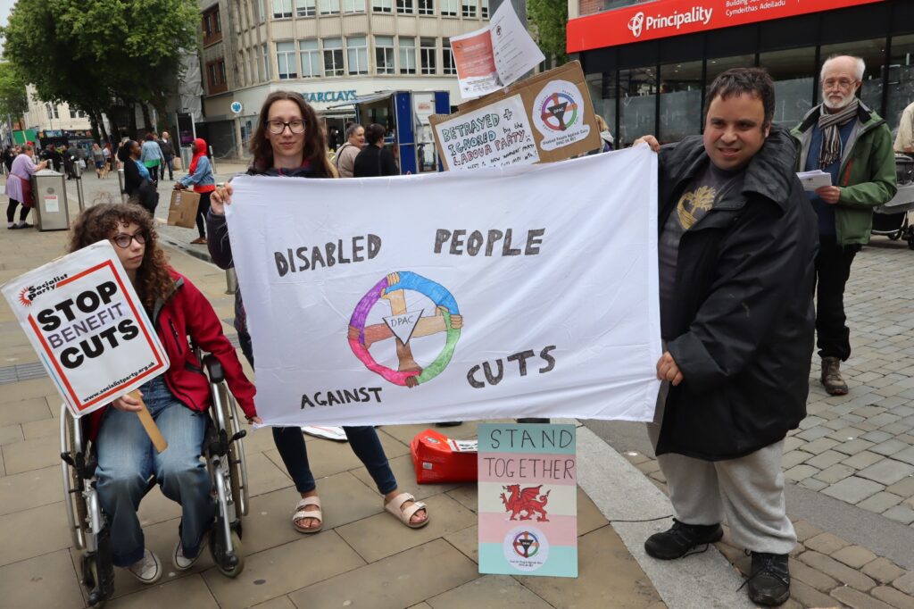 A photo of people protesting in Swansea city centre. They are holding a homemade banner that says "Disabled People Against Cuts" with the DPAC logo, which is a pink, purple, blue, green circle being held by four hands of varying skin tones. In the center is a triangle bearing the letters D P A C. Above the banner is a homemade placard that says "Betrayed and ignored by the Labour party". Beneath the banner is a placard in trans pride colours with a Welsh dragon and a DPAC logo. It says "Stand together". A young man and a young woman are holding the banner. To their left is a woman in a wheelchair holding a socialist party placard that says "stop benefit cuts". In the background is a man handing out DPAC leaflets.