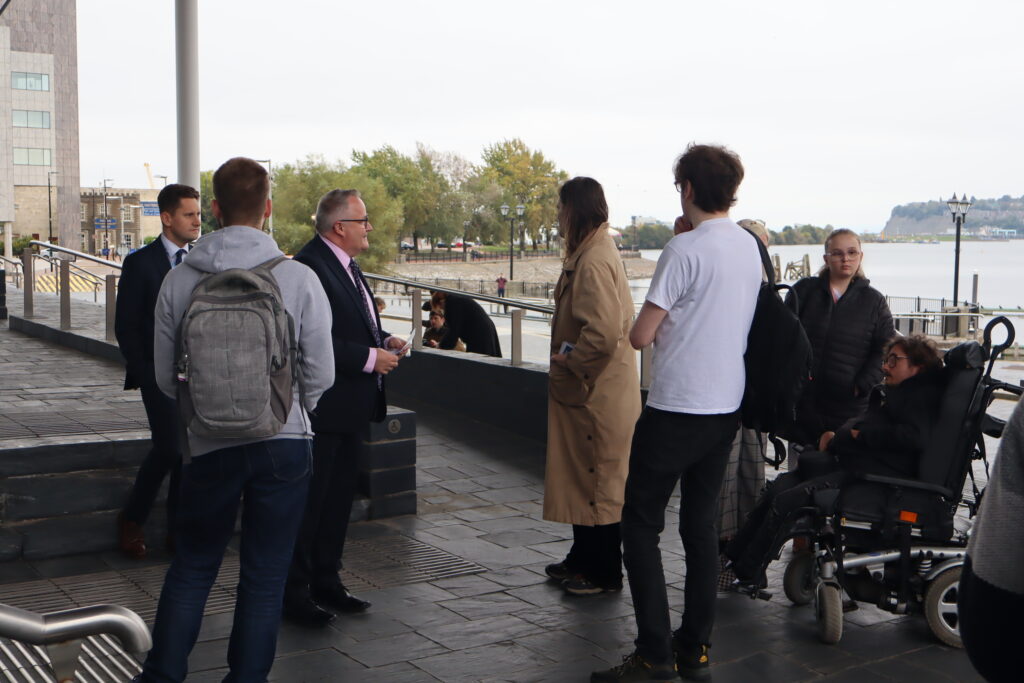 Darren Millar MS and staff converse with disability campaigners outside the Senedd. In the background are trees and the sea. It is picturesque, although overcast.