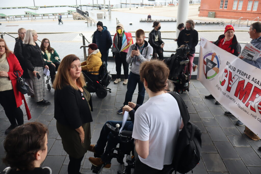 Sioned Williams MS talks to campaigners from Disabled People Against Cuts Cymru (DPAC Cymru) outside the Senedd. There are about 20 people in shot. Four people are users of powered wheelchairs. One person is wearing a yellow medical face mask and is draped in a Welsh flag with disability equality colours. Another wears a black medical face mask. People are standing around chatting, while Sioned is at the center of the photo. At the right of the photo is the DPAC Cymru banner being held by several people. The banner says "rights not charity." One person wears a hat that says PCS. One has a copy of the Socialist newspaper.