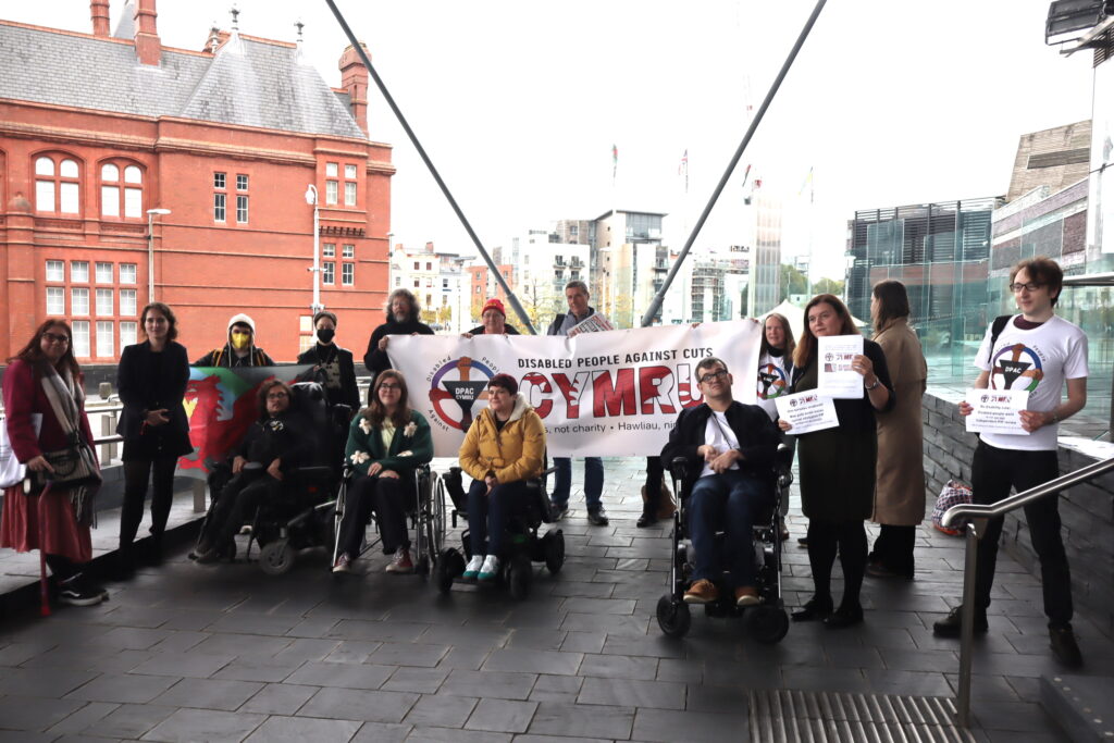 Disability campaign group Disabled People Against the Cuts Cymru pose for a group photo outside the Senedd (Welsh parliament) with Sioned Williams MS. Sioned is holding up the DPAC Cymru letter. There are 15 people in the photo. Two people are wearing DPAC t-shirts. Four people are using wheelchairs. Two people are wearing face-masks. One person is holding a flag showing a Welsh dragon with a disability equality colour background. Behind everyone is the DPAC Cymru banner. It is very large, and has five people holding it. It says Disabled People Against Cuts and then the word Cymru with a Welsh dragon background effect on the text. It has the DPAC logo which is a red, green, purple, blue wheel being held by four arms with different skin tones. At the center of the logo is an upside-down black triangle bearing the letters D P A C and the word Cymru. Behind the campaigners are various tall buildings in Cardiff. The nearest building is made of a striking orange brick.