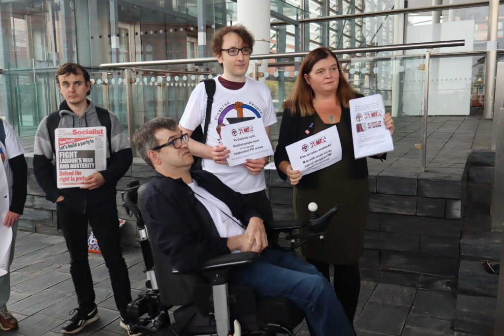 Lee Ellery and Ben Golightly from Disabled People Against Cuts Cymru (DPAC Cymru) pose for a photo with Sioned Williams. Lee Ellery is a wheelchair user and is in smart clothes. Ben is wearing a DPAC t-shirt. Ben is holding a sheet of paper with the DPAC Cymru logo that says "No disability cuts! Disabled people want to run our own independent PIP review!" Sioned is holding a sheet of paper with the same message translated into Welsh. She is also holding up the DPAC Cymru letter with 700 signatures. In the background, John Williams is standing to their leftwith a copy of a newspaper called the Socialist. On the cover of the newspaper, it says: "Your party: let's build a party to fight labour's war and austerity. defend the right to protest."