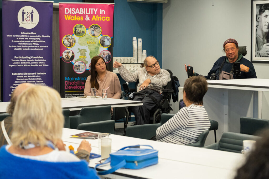 A photo of five people talking to each other indoors. Two of the people are wheelchair users. In the background is a banner that has the title "disability in Wales and Africa"